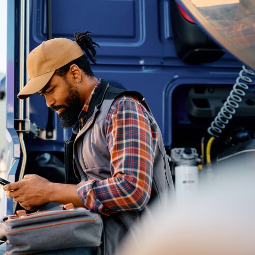 African American professional driver using smart phone on truck parking lot.